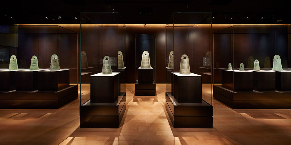 国宝「桜ヶ丘銅鐸・銅戈」を一堂に展示する2階『コレクション展示室』。茶褐色の銅板を背景に銅鐸が整然と並び、格調高く落ち着いた鑑賞空間となっている。