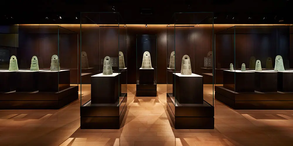 The second floor "Collection Exhibition Room" displays the national treasure "Sakuragaoka Bronze Bells and Bronze Spears" all in one place. The bronze bells are lined up neatly against the background of brown copper plates, creating a dignified and calm viewing space.