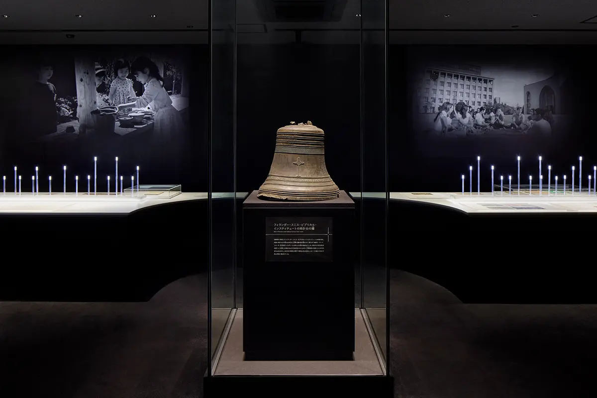 History of Aoyama Gakuin ③: Light of the Trajectory | At the beginning of the room, the bell from the clock tower of the school building, which was completed in 1886, is displayed as a symbol of the school's history. In the background, slides of the school's current appearance are projected, creating an exhibition scene that connects the past and present.