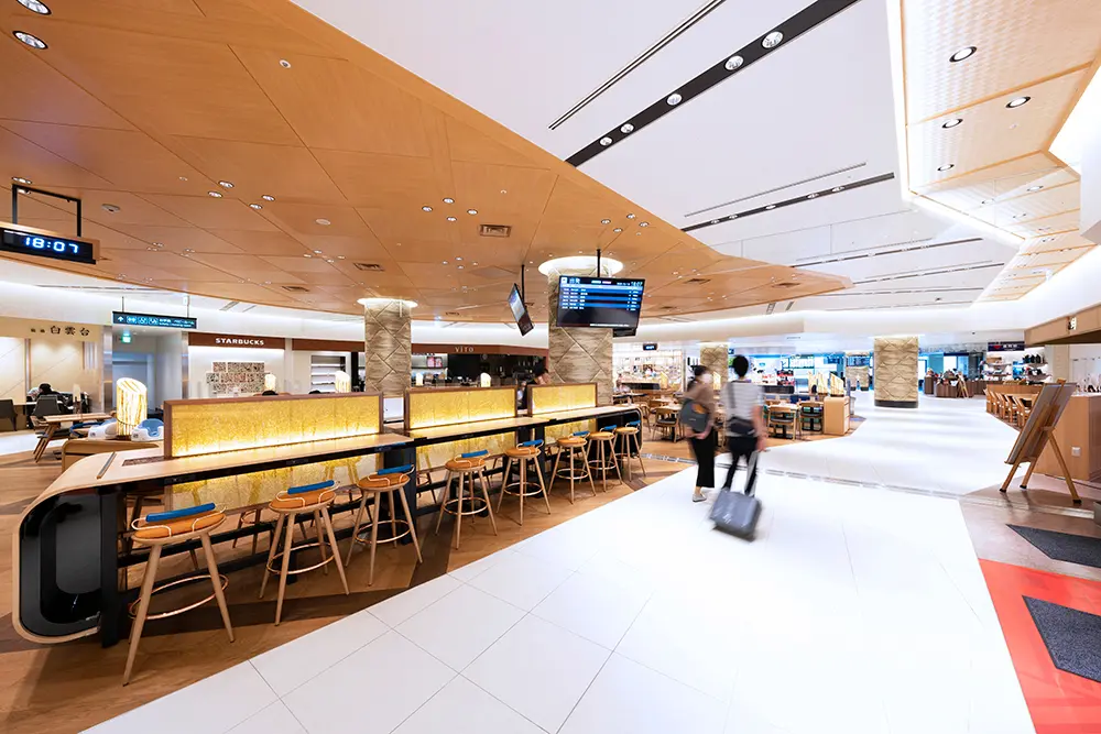 An open space adjacent to the passageway leading to the boarding gate. It is used as a shared seating area for the surrounding stores, for eating and drinking, resting, and working.